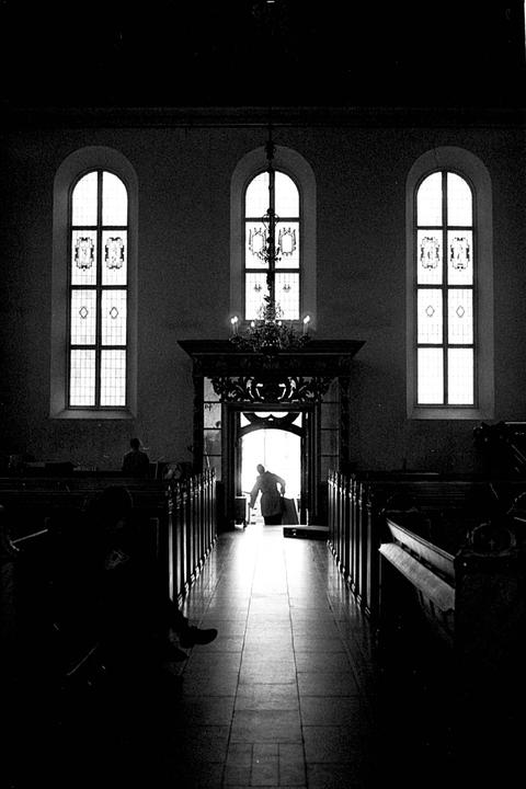 A person enters a church through a brightly lit doorway, with rows of pews on either side and three tall arched windows above.
