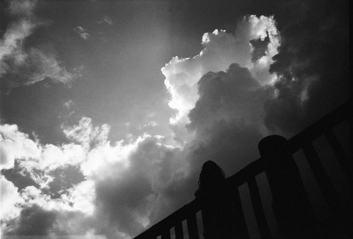 A woman stands silhouetted against a cloudy sky, looking out from behind a railing.