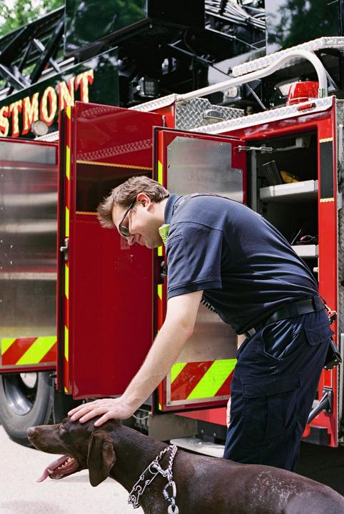 A man wearing sunglasses and a dark blue uniform pets a brown dog with a chain collar next to a red fire truck.