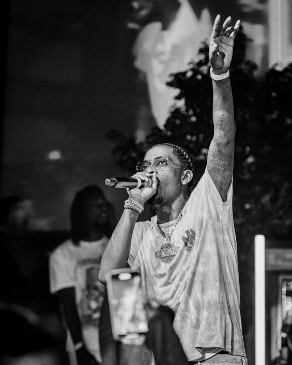 A man with braided hair and glasses holds a microphone and raises his arm while performing on stage.
