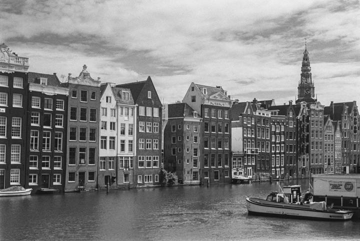 A canal boat floats on a canal lined with tall, narrow buildings and a clock tower under a cloudy sky.