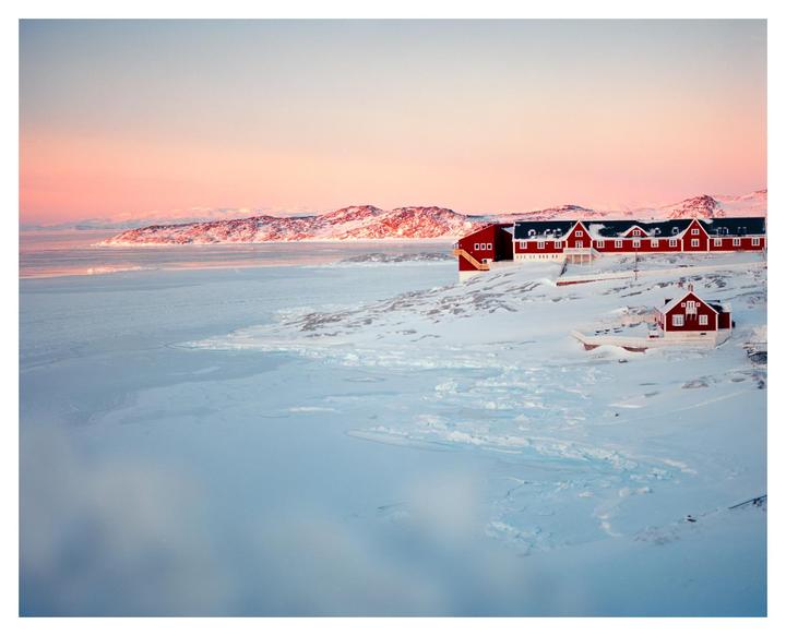 Red houses sit on a snow-covered landscape next to a frozen body of water with mountains in the distance under a pink sky.