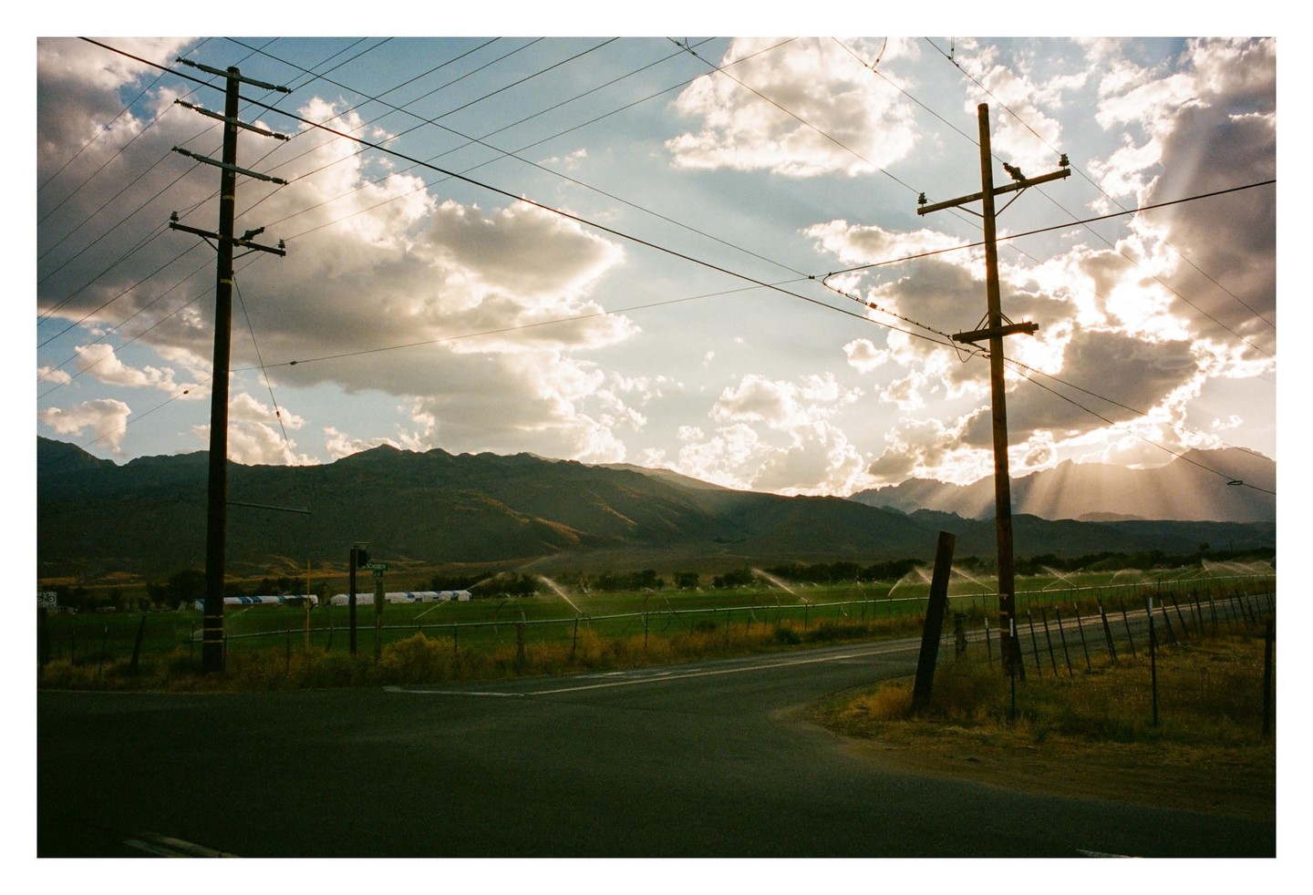 A road curves past a field with sprinklers and power lines under a cloudy sky with sun rays over mountains.