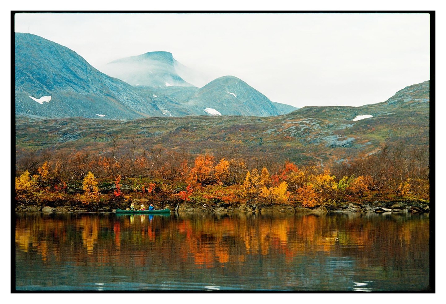 A canoe with three people floats on a lake surrounded by autumn trees and mountains.