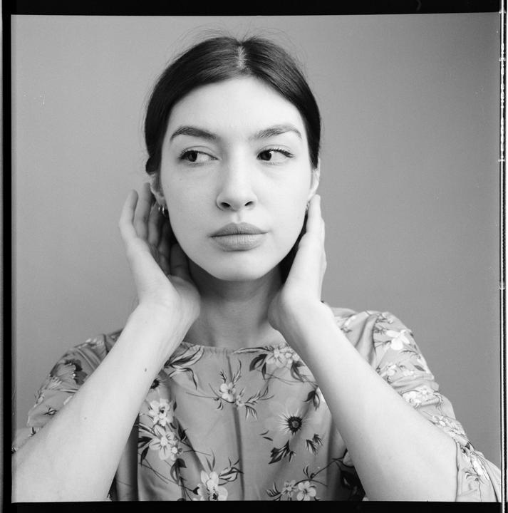 A woman with dark hair and a floral top holds her hands to her ears and looks to the side.