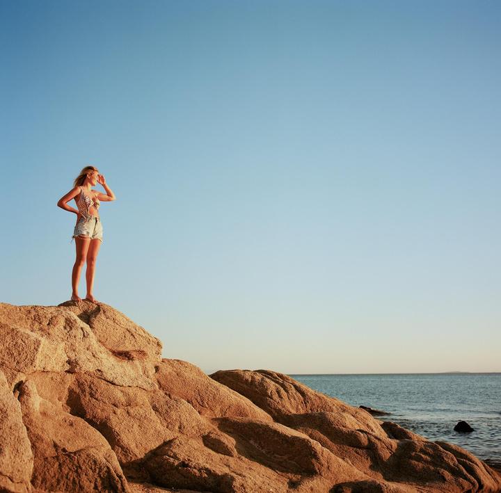 A woman stands on a rocky outcrop overlooking the ocean under a clear sky.