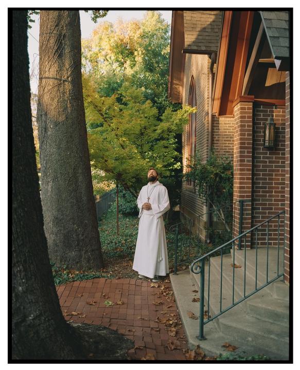 A man in a white robe stands on a brick path next to a building with trees in the background.