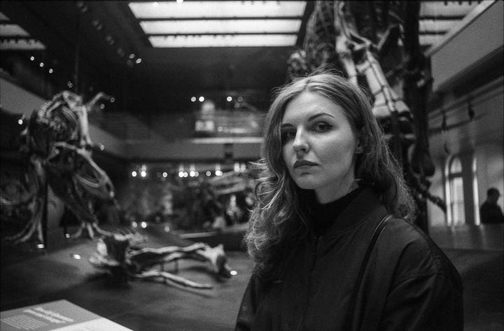 A woman with long hair looks at the viewer in a museum with dinosaur skeletons in the background.