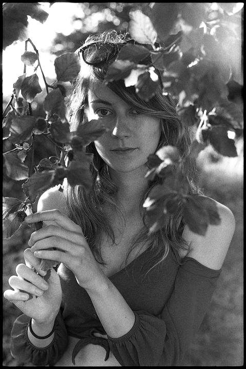 A woman with long hair and sunglasses on her head looks directly at the viewer through a curtain of leaves.