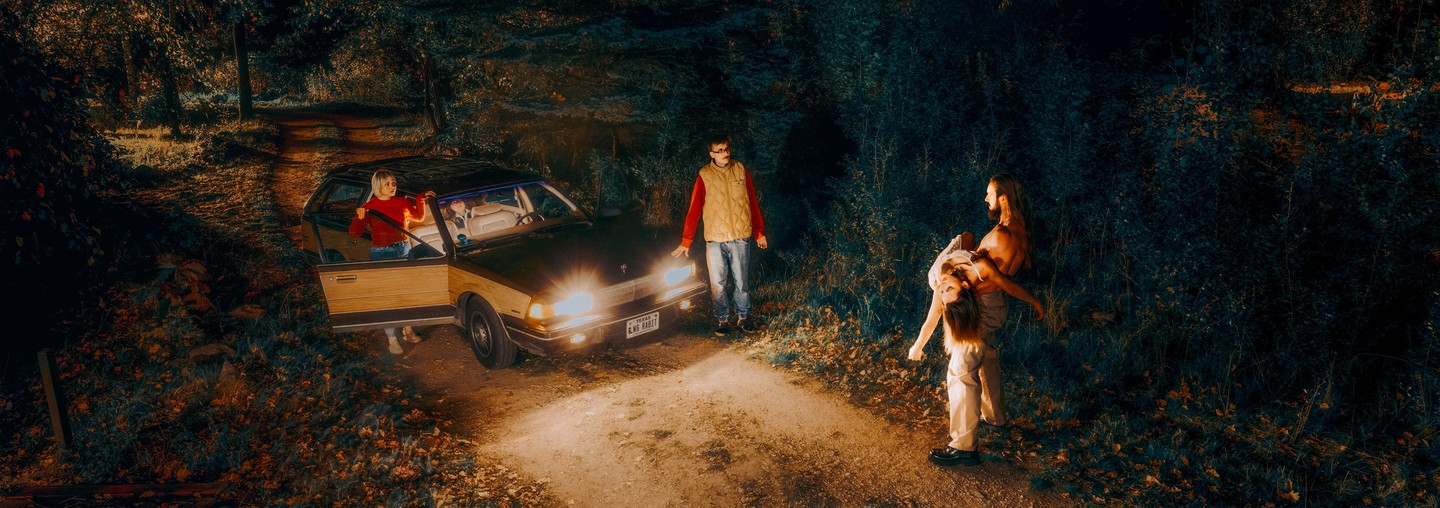 A woman exits a car with its headlights on, while a man stands by the front of the car and another man carries a woman in a dark, wooded area at night.