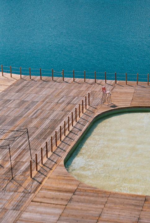 A wooden deck with a small pool and a single red chair overlooks a large body of blue water.
