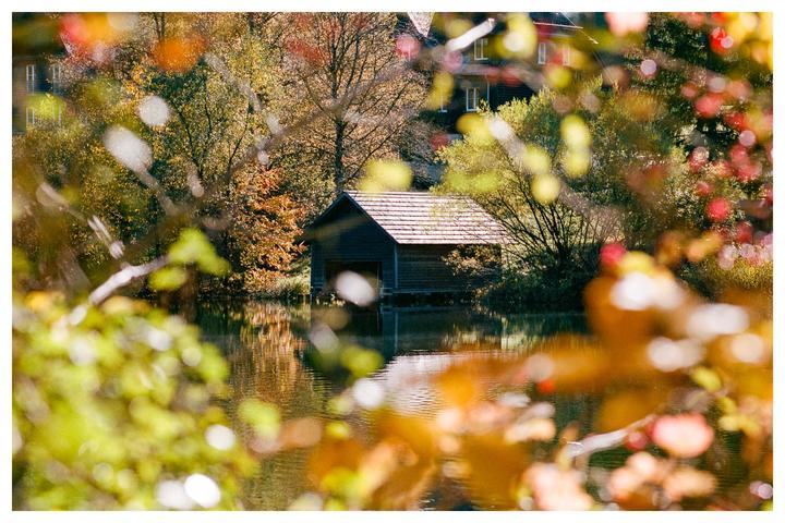 A wooden boathouse with a shingled roof sits on a lake surrounded by autumn foliage.