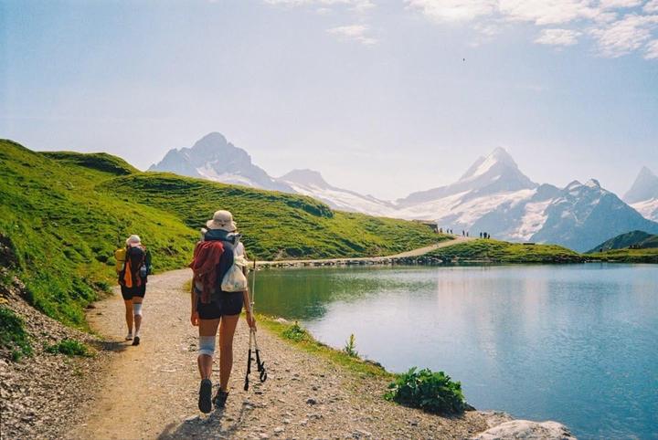 Two hikers walk along a path next to a lake with snow-capped mountains in the distance.