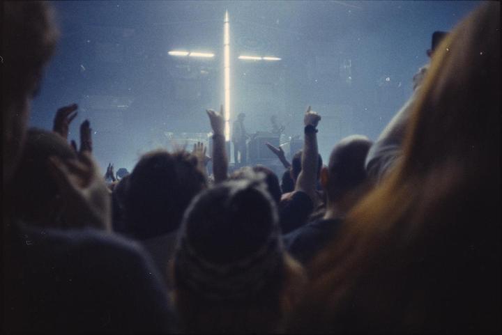 A crowd of people with their hands raised watches a band perform on a stage lit by a cross-shaped light.