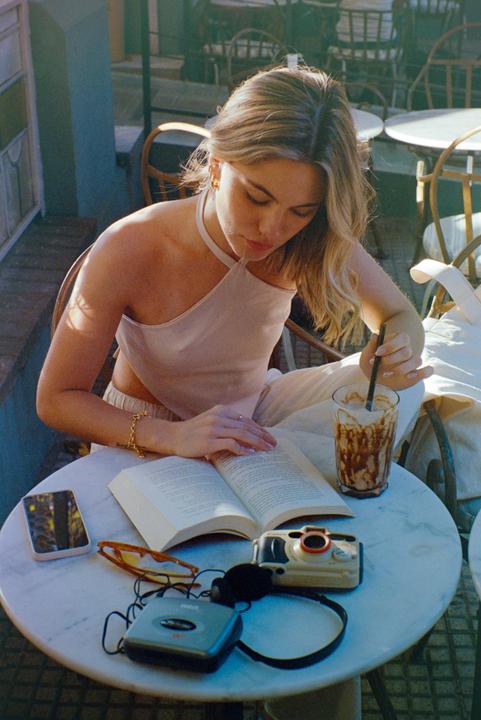 A woman reads a book at an outdoor cafe table with a drink, a phone, sunglasses, a camera, and a cassette player.