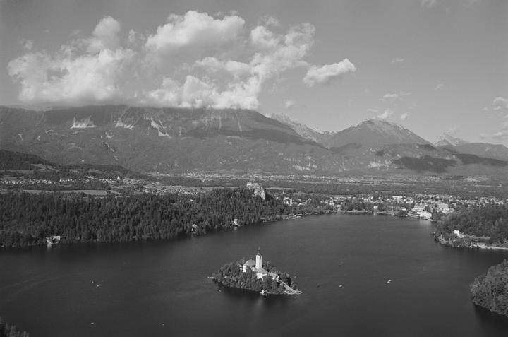 A small island with a church and bell tower sits in a lake surrounded by a town, forest, and mountains under a cloudy sky.