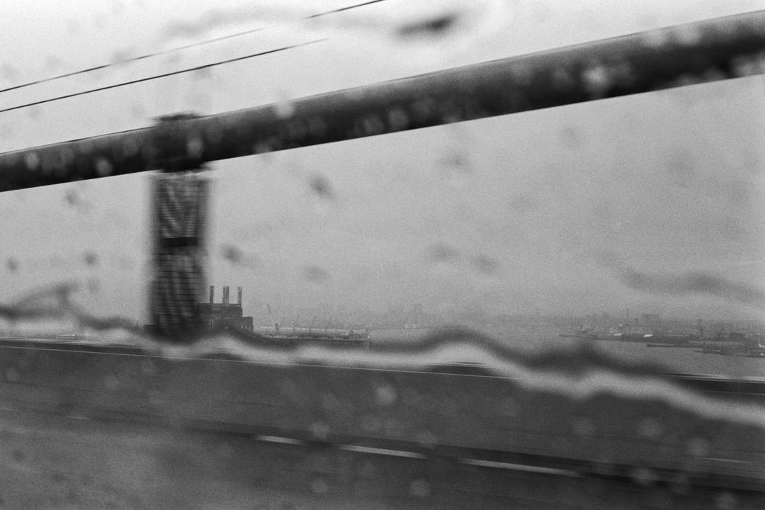 A rainy view from a moving vehicle shows a bridge railing, a distant industrial building, and a body of water with a city skyline on the horizon.