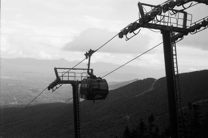 A gondola car hangs from a cable above a mountain landscape with forested slopes and distant peaks under a cloudy sky.