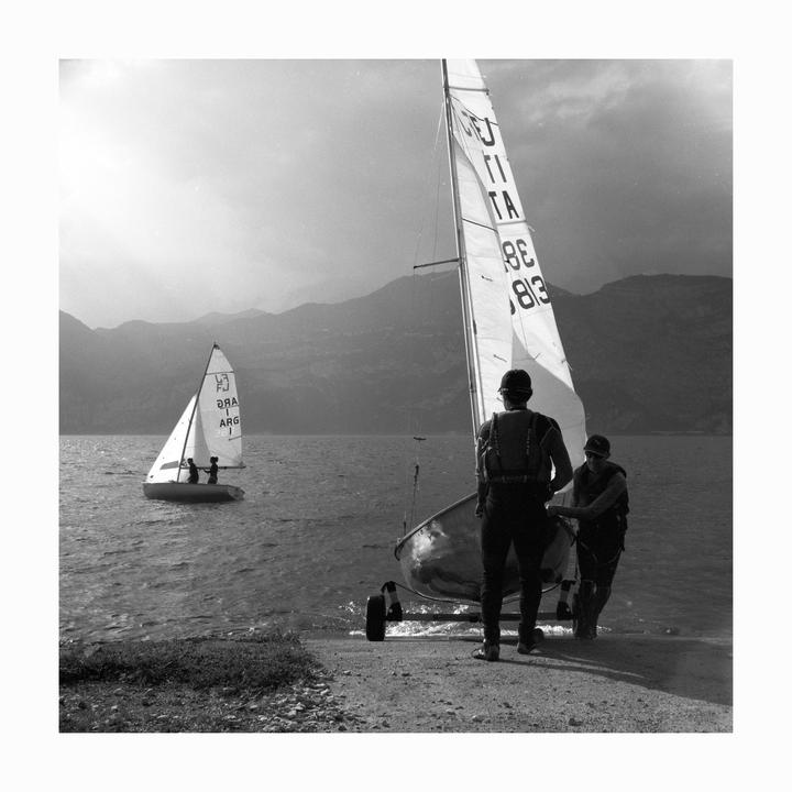 Two men launch a sailboat from a rocky shore into a lake with mountains in the background.