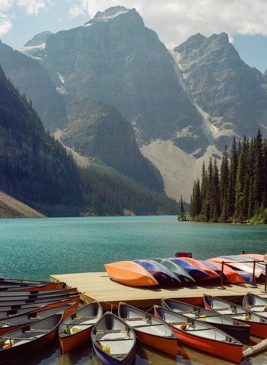 Colorful canoes and kayaks are docked on a wooden pier on a turquoise lake surrounded by mountains and pine trees.