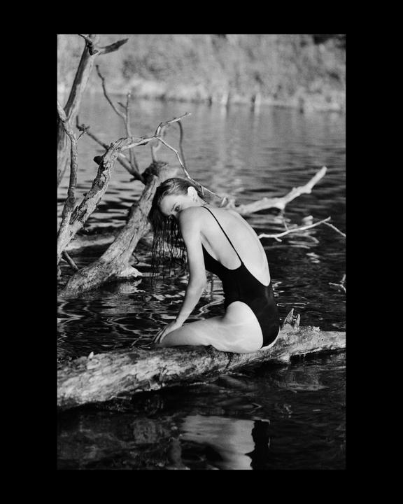 A woman in a swimsuit sits on a fallen tree branch in the water.