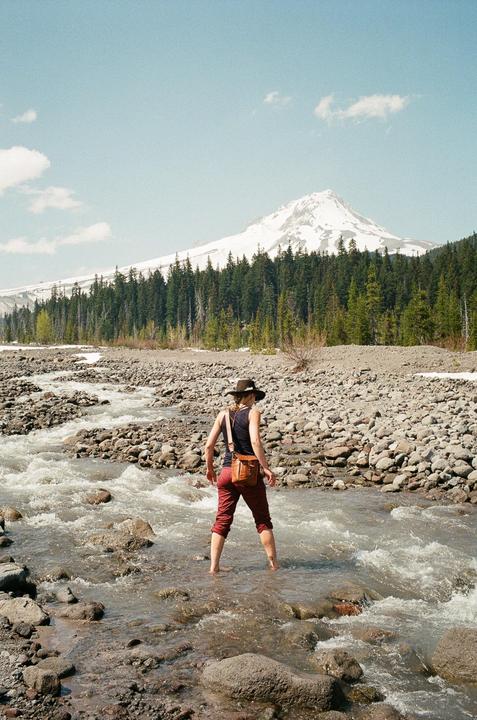 A woman stands barefoot in a rocky river with a snow-capped mountain and forest in the background.