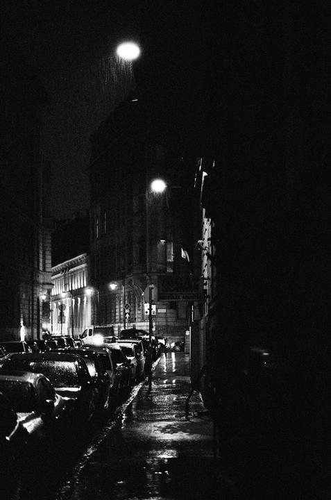 A dark street is lined with parked cars and illuminated by streetlights, with rain visible in the light.
