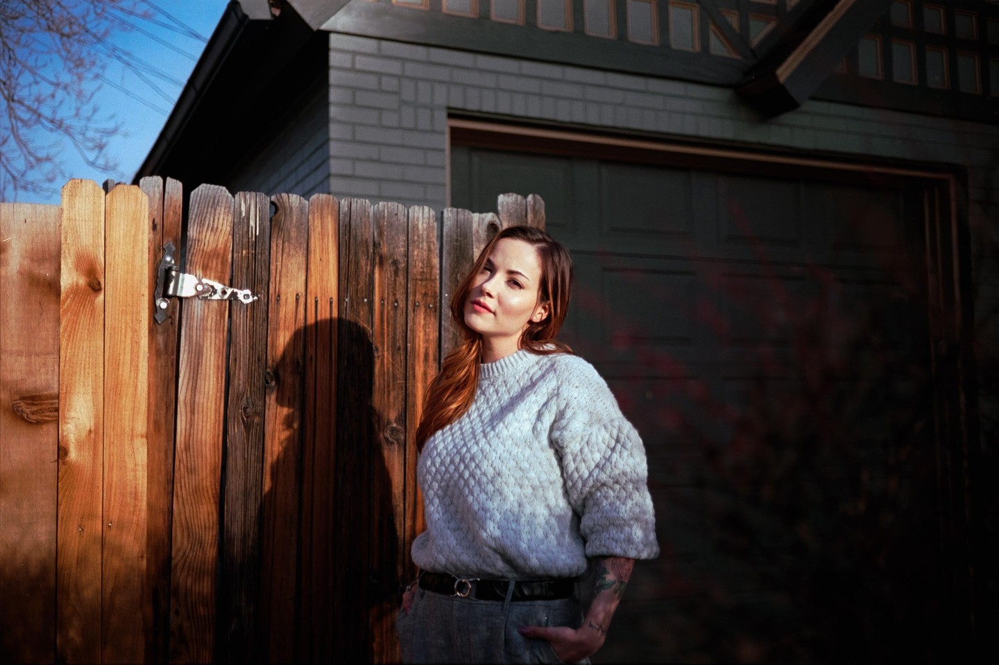 A woman with long hair and a light sweater stands in front of a wooden fence with a shadow of her head and shoulders cast upon it.