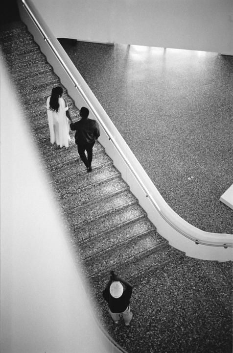 A couple walks down a curved staircase while a man stands below, looking up.