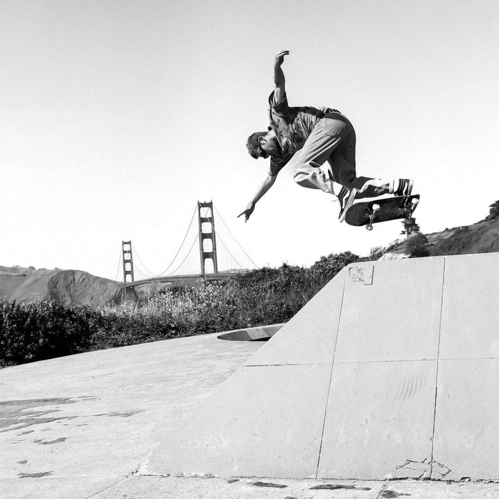 A man on a skateboard is captured mid-air above a concrete ramp with the Golden Gate Bridge visible in the background.