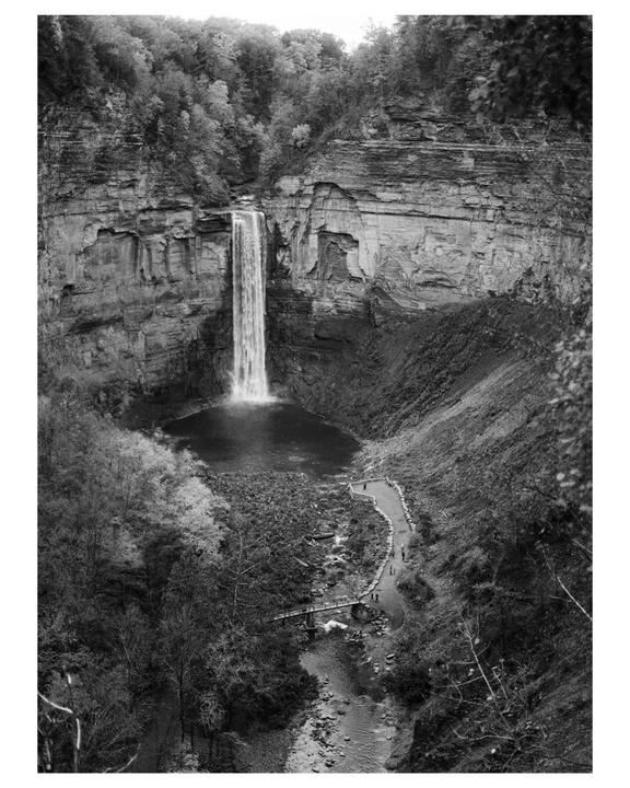 A tall waterfall cascades into a pool at the base of a cliff, with a path and bridge visible below.