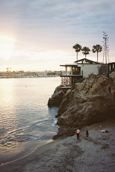 A modern house with palm trees sits on a rocky cliff overlooking a bay with a sandy beach where a couple is relaxing.