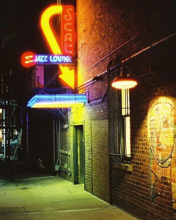 A neon sign for a jazz lounge illuminates a brick alleyway at night.
