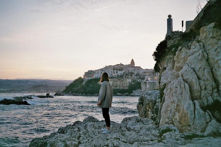 A woman stands on a rocky shore looking out at a town built on a cliff overlooking the sea at dusk.