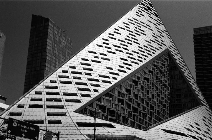 A triangular building with many windows is reflected in another building, with a tall rectangular building in the background.
