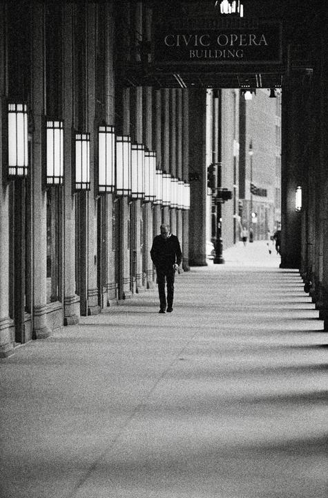 A man walks down a long, columned walkway with many illuminated lamps on the left and a sign for the Civic Opera Building overhead.