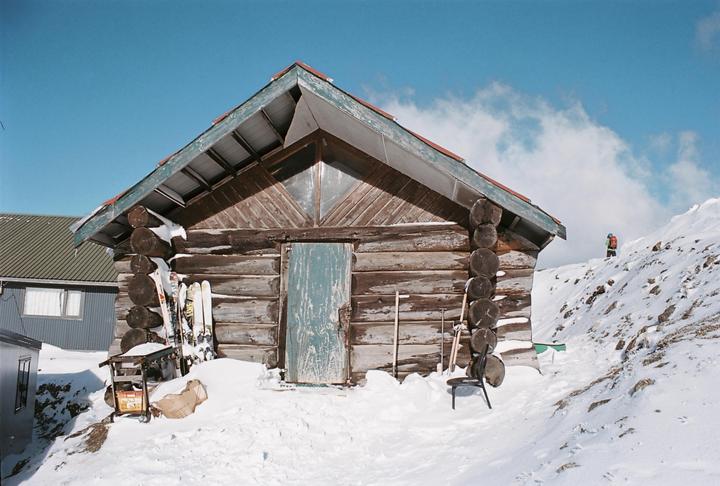 A wooden cabin with skis leaning against it stands in a snowy landscape under a blue sky, with a person visible on a distant snowy hill.