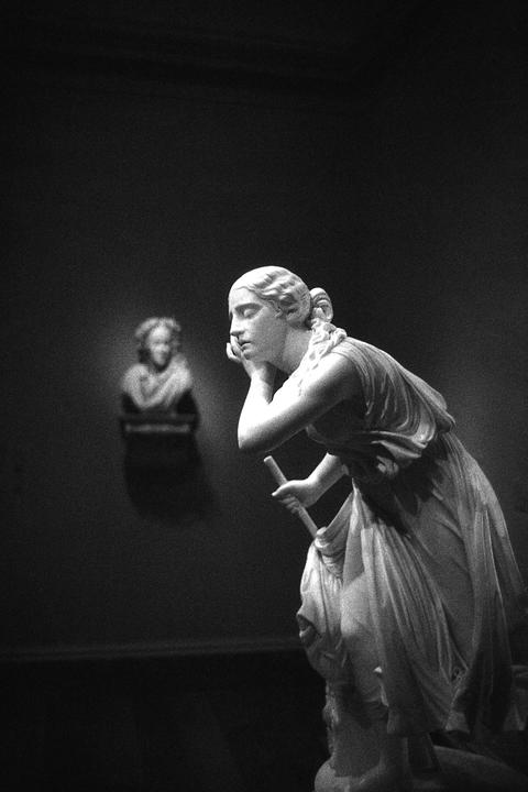 A white marble statue of a woman with her hand to her face and holding a staff stands in front of a blurred bust on a dark wall.