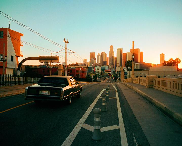 A black car drives on a street towards a city skyline at sunset.