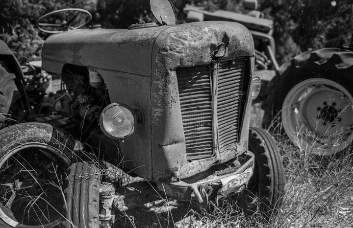 A black and white close-up shows the front of an old, rusty tractor with a single headlight and a damaged grille, surrounded by tall grass.