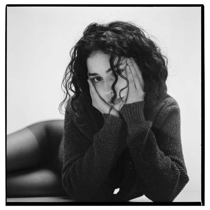 A woman with curly hair lies on her side, looking at the viewer with her hands cupping her face.