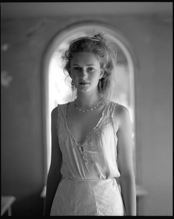A woman with curly hair and a pearl necklace stands in front of an arched doorway.