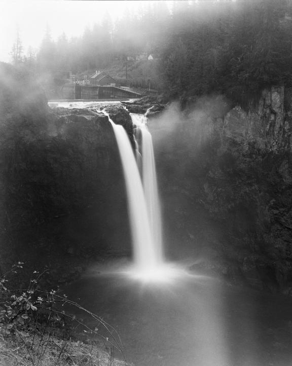 A long exposure captures a waterfall cascading into a pool below, with a building and forest visible through mist in the background.