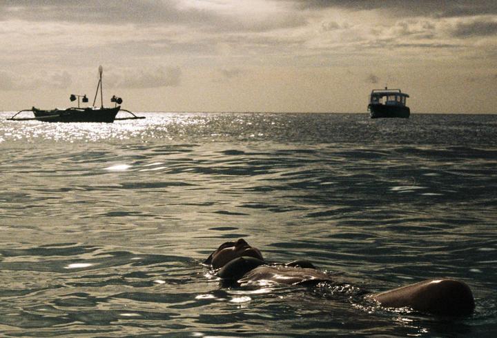 A woman floats on her back in the ocean with two boats in the distance under a cloudy sky.