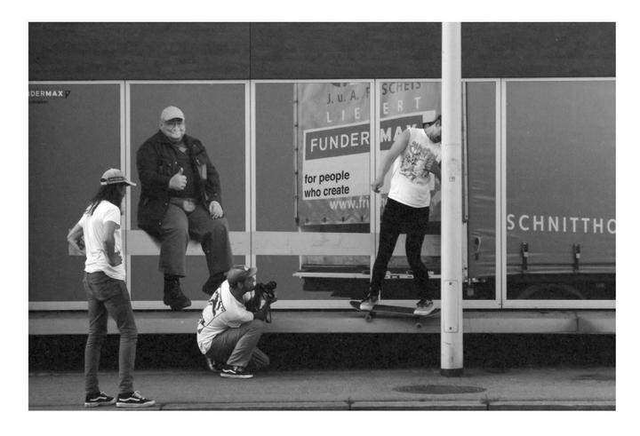 Four men are gathered on a street next to a building with large windows, one of whom is on a skateboard and another is holding a camera.
