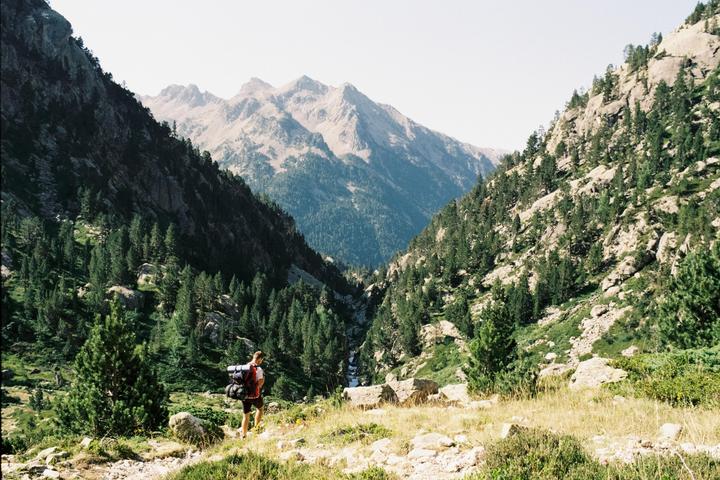 A man with a backpack stands on a grassy hill overlooking a mountain valley with a river.