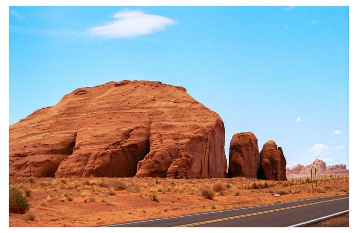 A road runs through a desert landscape with large red rock formations under a bright blue sky.