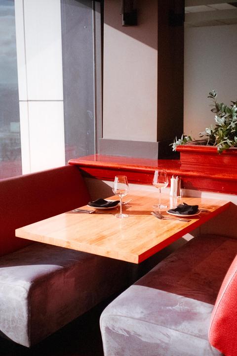 A restaurant booth with a wooden table set for two is illuminated by sunlight streaming through a large window.