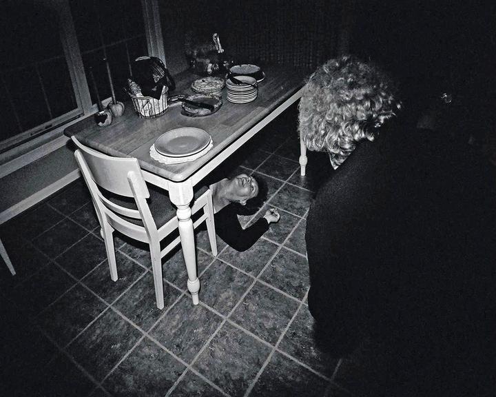A woman lies on the floor under a dining table while another person stands nearby.