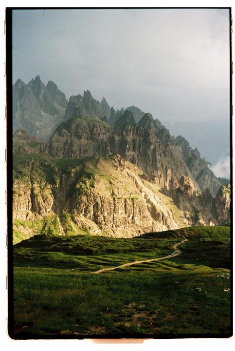 A winding path leads through a green, grassy field towards a range of jagged, rocky mountains under a hazy sky.